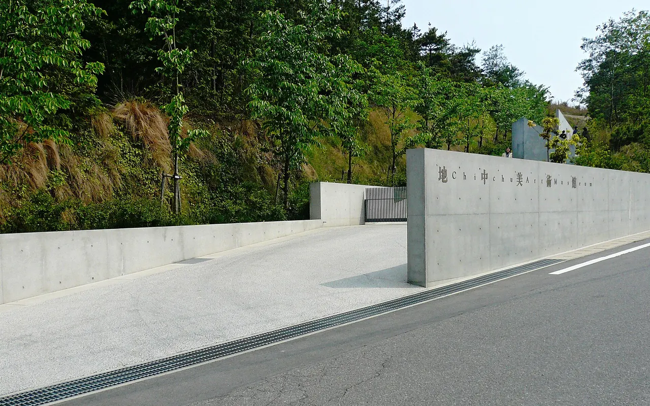 Eine lange Betonmauer an der Straße mit dem Namen des Chichu Art Museum, dahinter Bäume